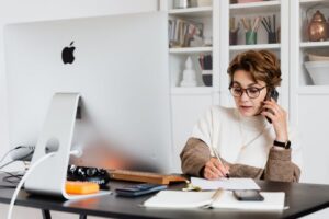 person-at-desk-with-monitor-talking-on-phone-and-writing-notes-on-notepad